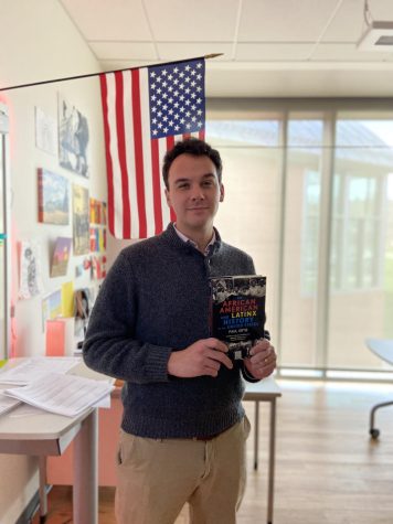 Mr. Ames poses with a book he reads under an American flag in his classroom
