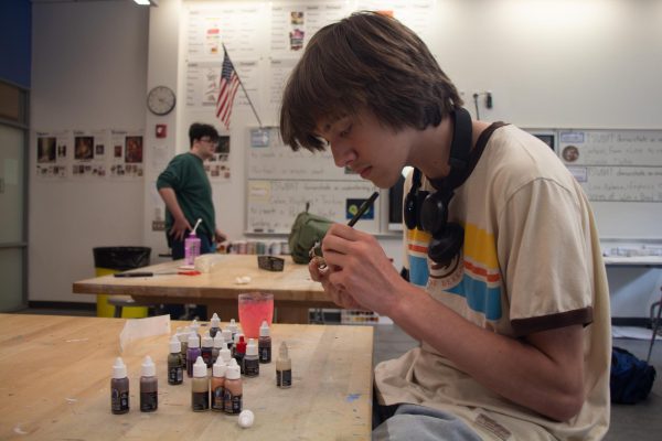 A student focusing on painting a small figurine, with various small paint bottles on the table infront of him.