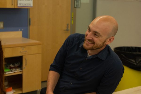 Justin Sylvia, the Morgan School art teacher, sitting and smiling while conversing with other students.